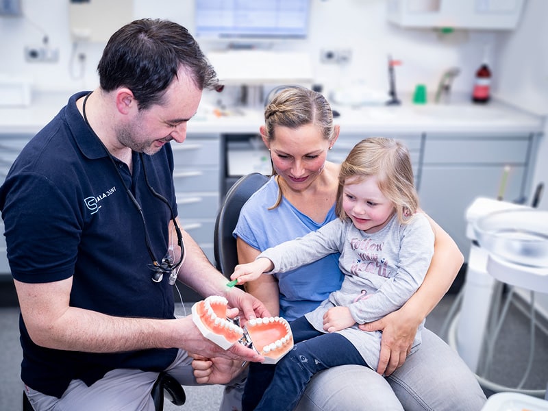 Dr. Christian Lichthardt erklärt einem Kind auf dem Schoß der Mutter wie man richtig Zähne putzt